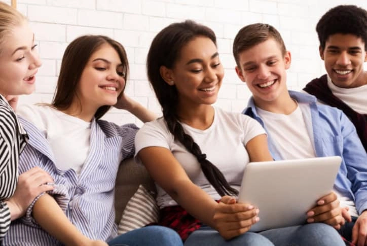 Grupa nasmejanih devojaka i momaka gledaju nešto na tabletu