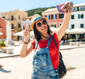 Devojka se naočarima i šeširom se slika selfi i drži dva prsta podignuta