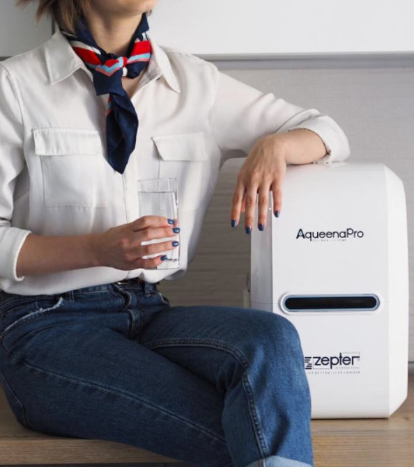 Žena sa čašom vode u ruci sedi pored prečišćivača vode AqueenaPro